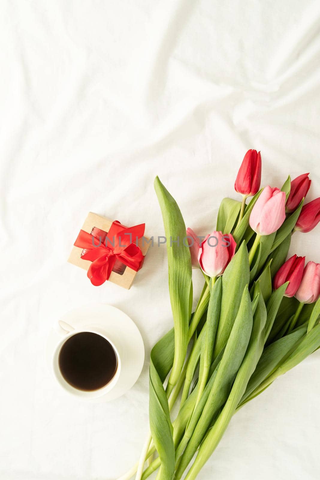 Holiday and celebration. Top view of pink tulips, coffee cup and gift box on white bed, flat lay