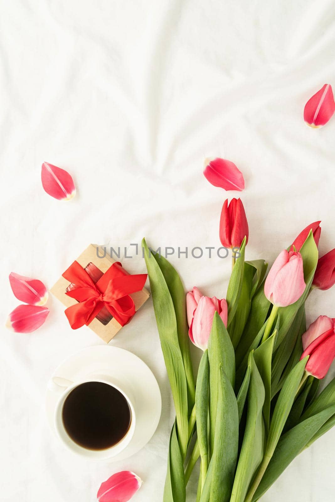 Holiday and celebration. Top view of pink tulips, coffee cup and gift box on white bed, flat lay