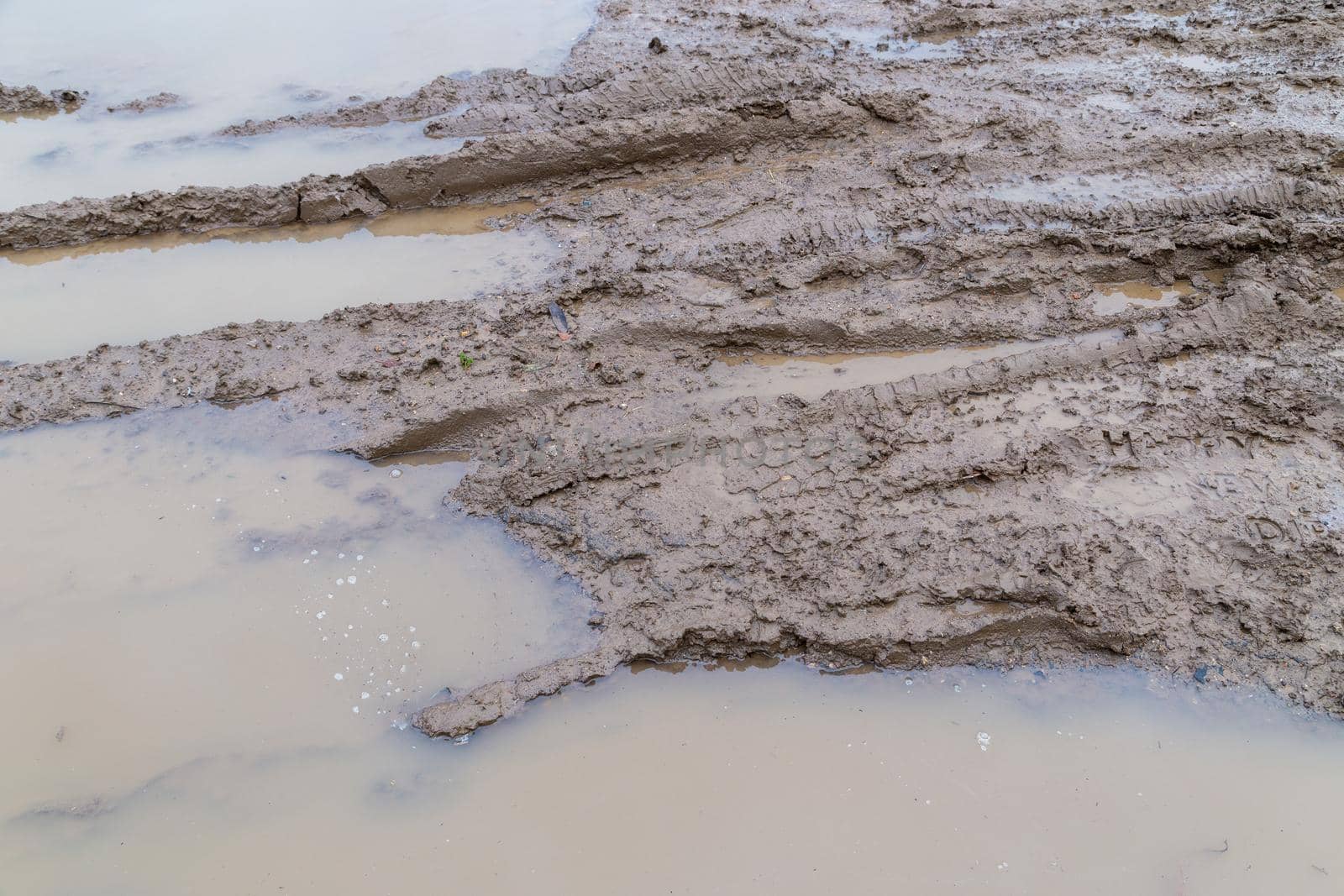dirty wet clay mud road with tire tracks and puddles at day time - full frame background with hard sun light