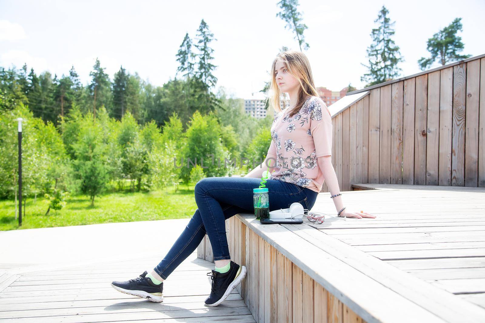 A young girl of 20 years old Caucasian appearance enjoys the sun and weather while sitting on a wooden podium in the park on a summer day.The girl is dressed in a floral T-shirt and jeans