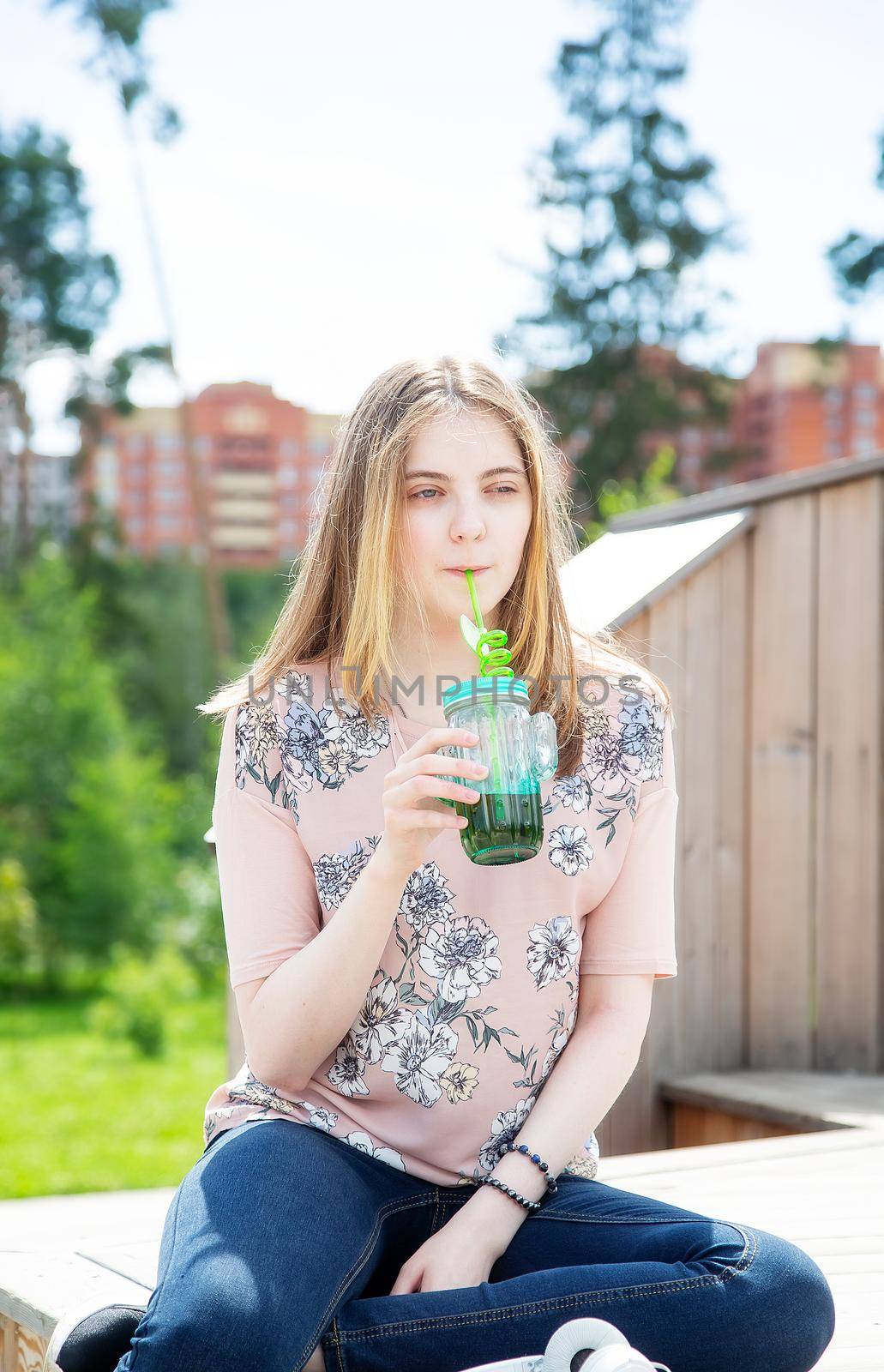 A young girl of 20 years old Caucasian appearance enjoys the sun and weather and drinks smoothies from a large glass while sitting on a wooden podium in the park on a summer day.