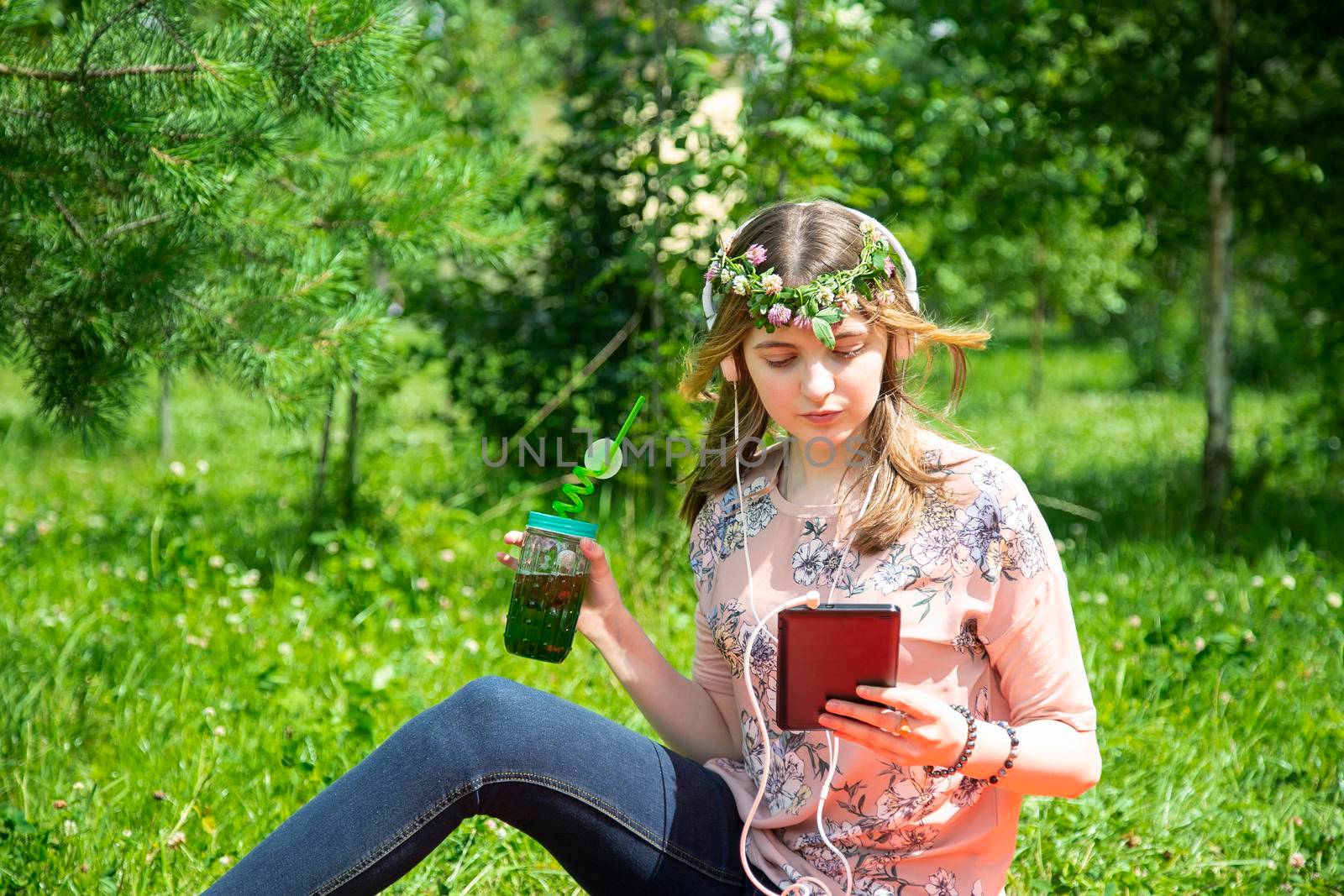 A young girl of 20 years old Caucasian appearance listens to music on her mobile phone and drinks smoothies from a large glass, sitting on the lawn in the park on a summer day.