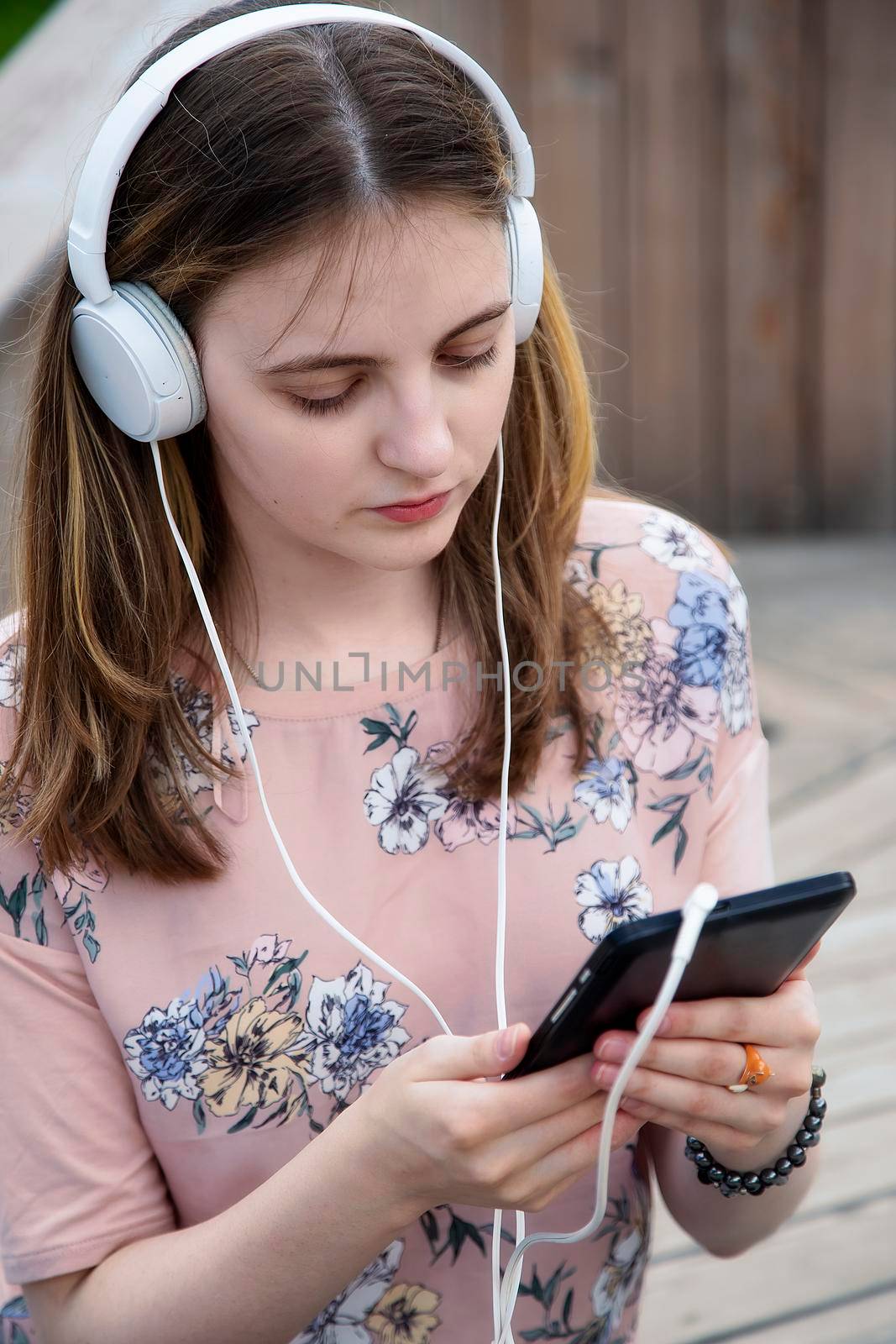 A young girl 20 years old Caucasian appearance looks into her mobile phone, writes a text message while sitting on a wooden podium in the park on a summer day.The girl is dressed in a floral T-shirt 