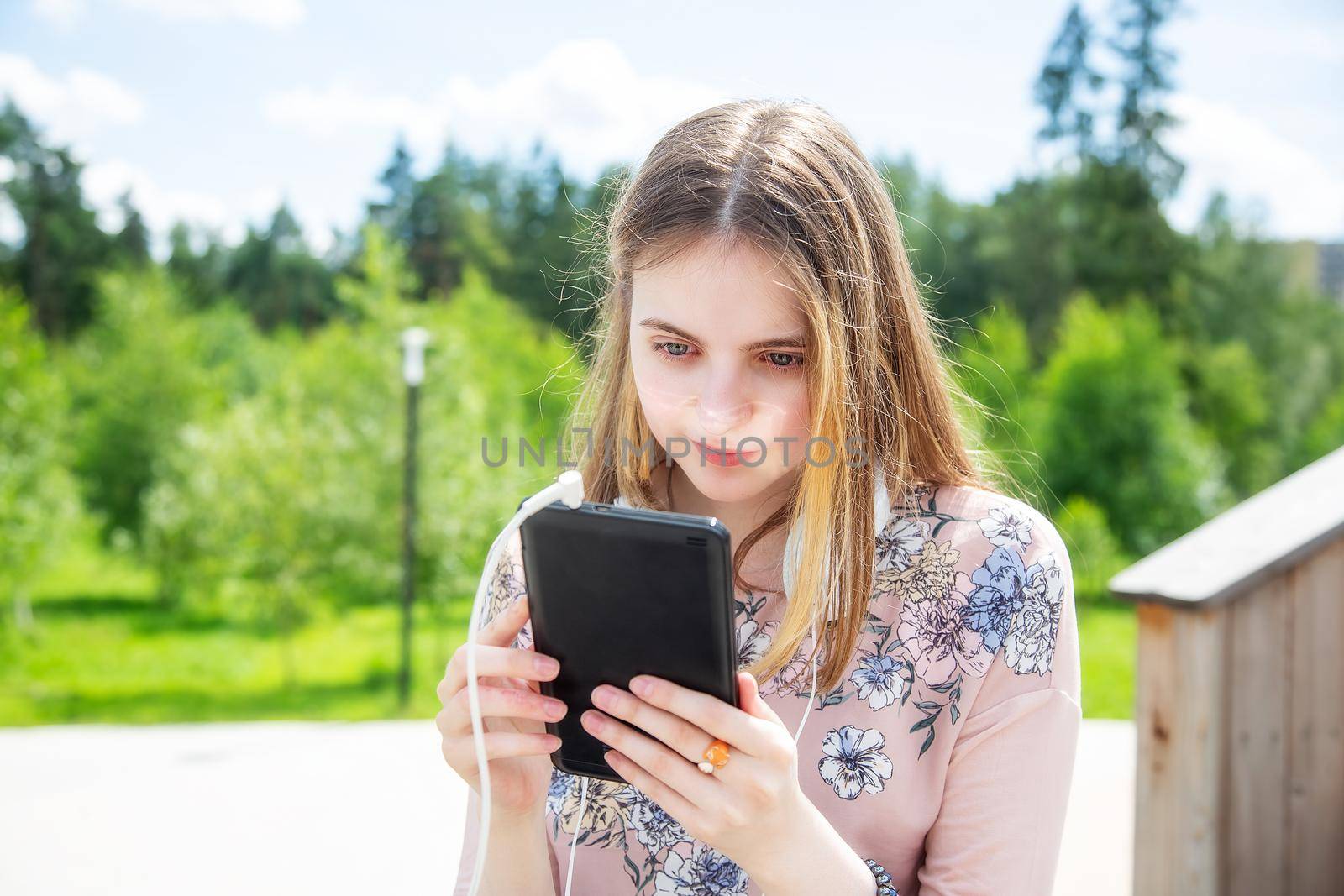 A young girl 20 years old Caucasian appearance looks into her mobile phone, writes a text message while sitting on a wooden podium in the park on a summer day.The girl is dressed in a floral T-shirt