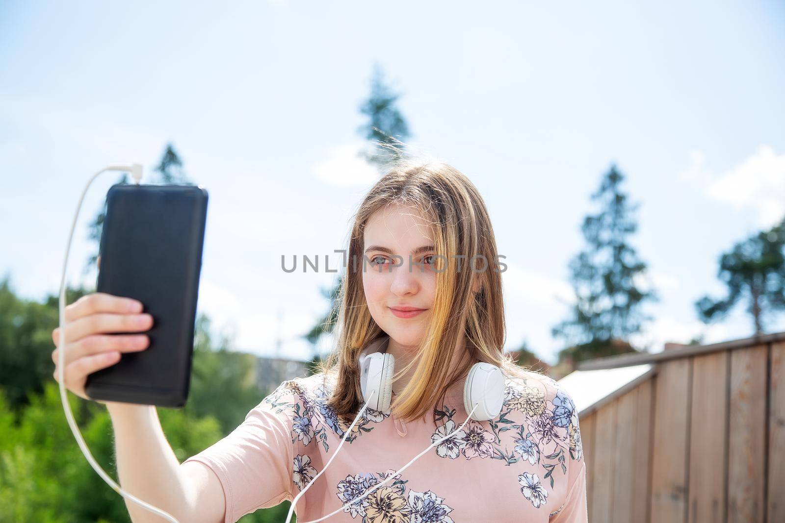 A young girl of 20 years old Caucasian appearance makes a selfie on her mobile phone while sitting on a wooden podium in the park on a summer day.The girl is dressed in a floral T-shirt and jeans, 