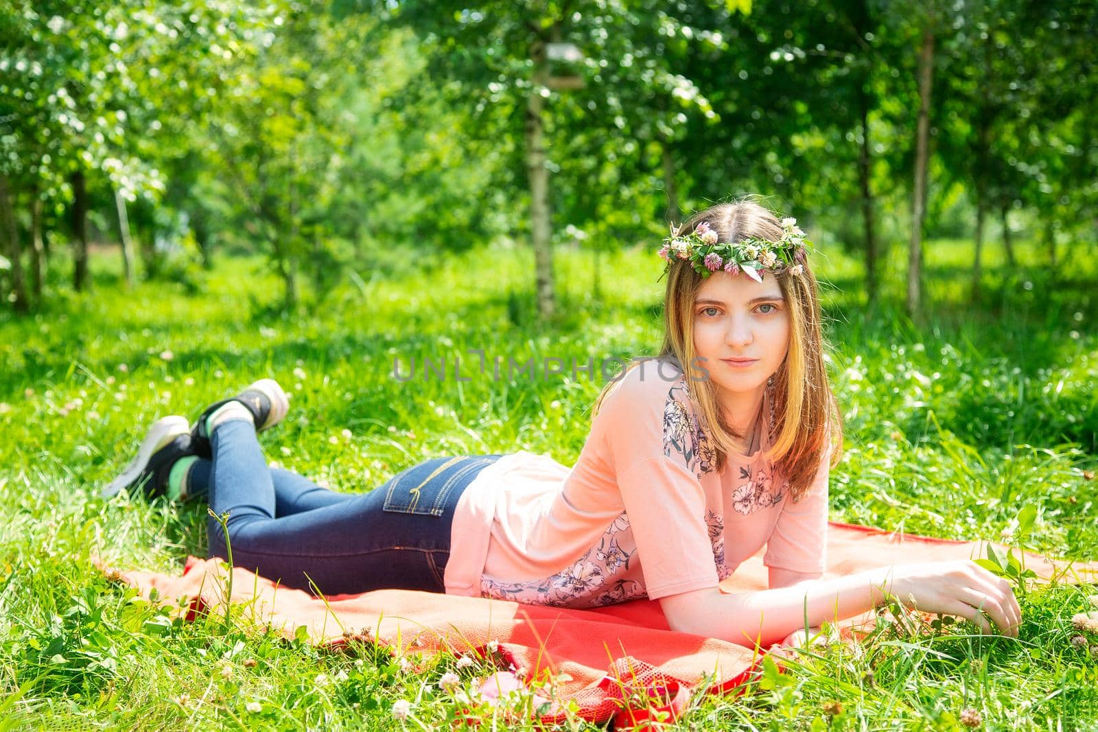 Young girl 20 years old Caucasian appearance smiling looking at the camera while lying on the lawn in the park on a summer day. The girl is dressed in a T-shirt and jeans and a wreath of wildflowers.