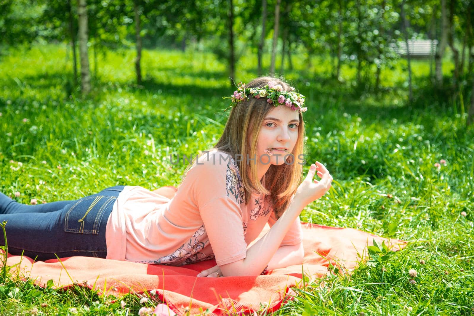 Young girl 20 years old Caucasian appearance smiling looking at the camera while lying on the lawn in the park on a summer day. The girl is dressed in a T-shirt and jeans and a wreath of wildflowers.