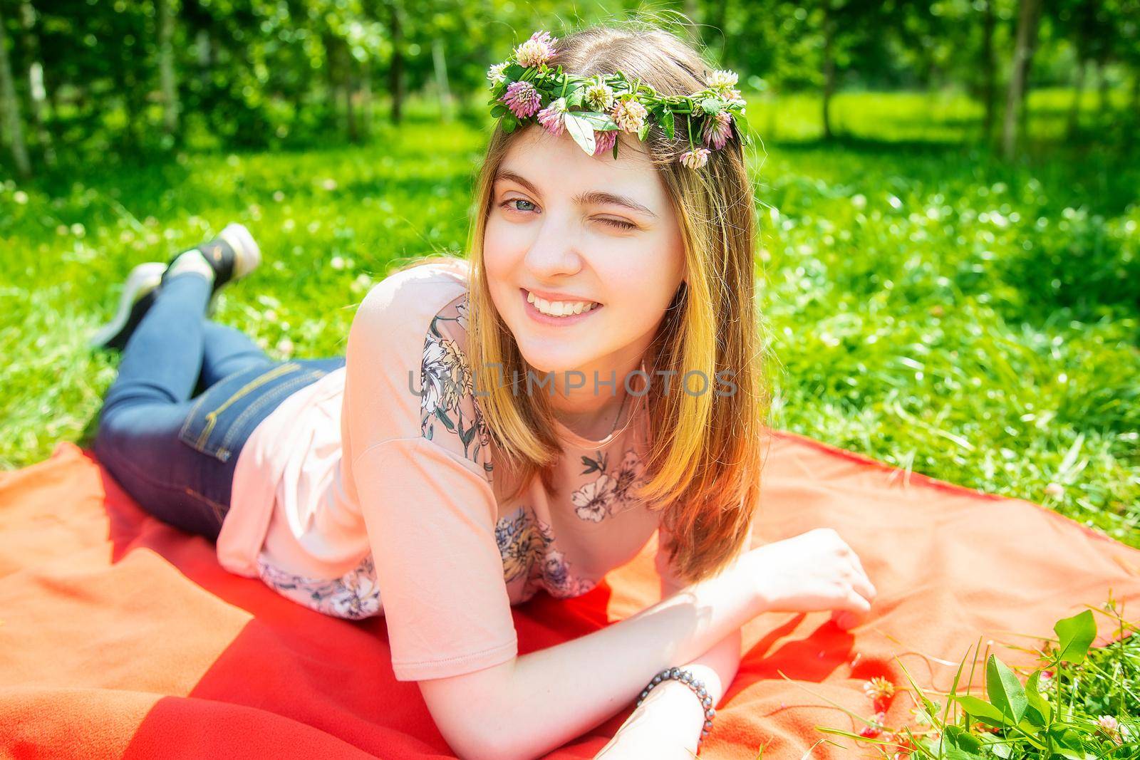 Young girl 20 years old Caucasian appearance smiling looking at the camera while lying on the lawn in the park on a summer day. The girl is dressed in a T-shirt and jeans and a wreath of wildflowers.