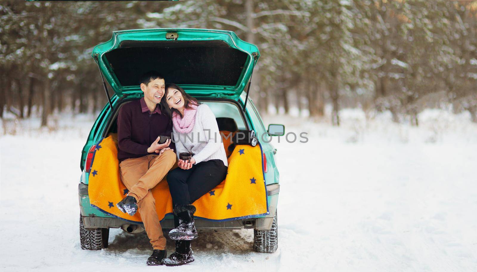 Happy couple sitting in the open trunk of a car in winter outdoors with mugs of hot tea in their hands