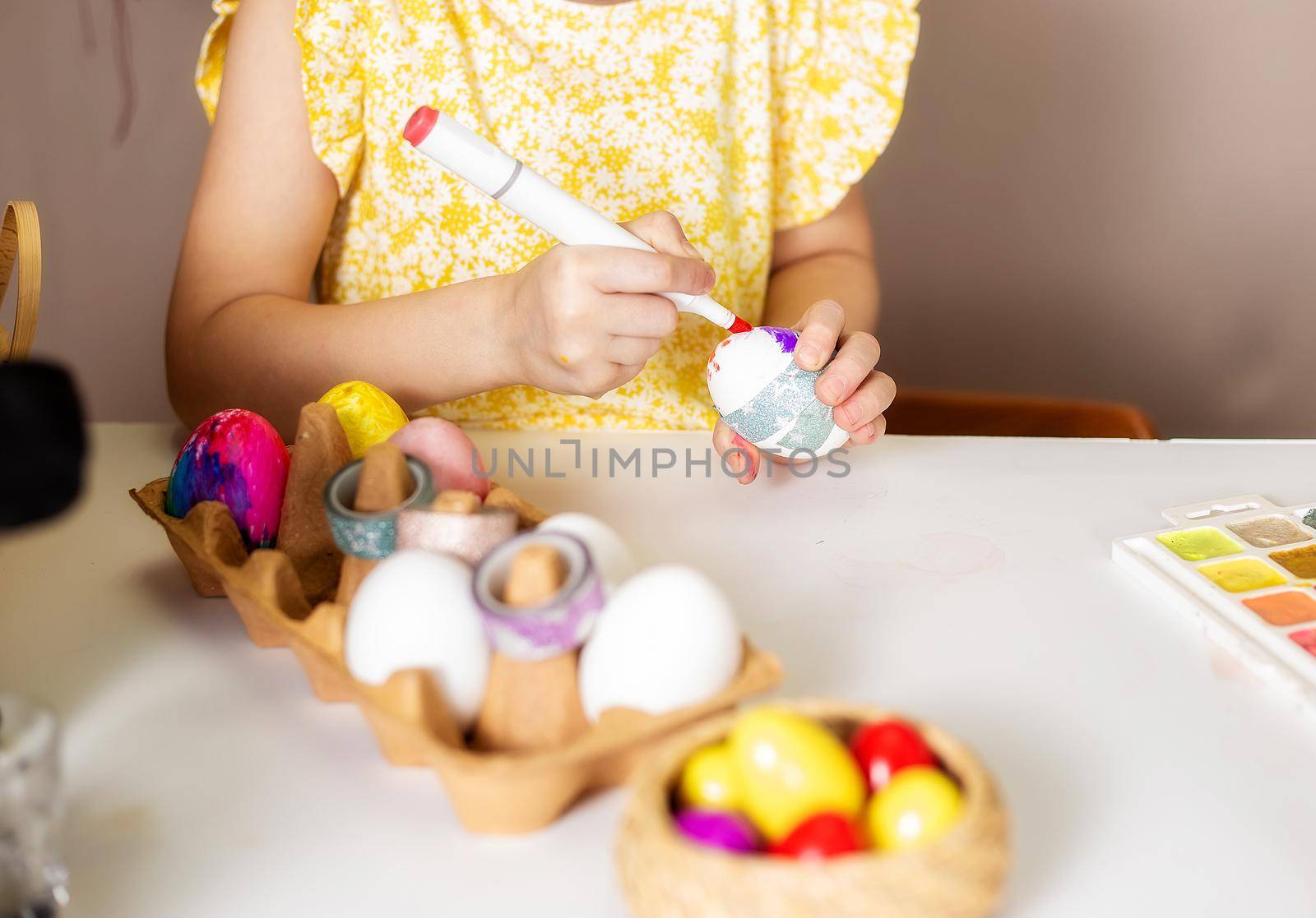 A close-up on the hands of a small Caucasian girl 5 years old paints eggs with special markers paints for the Christian spring holiday of Easter. Girl dressed in a yellow floral dress