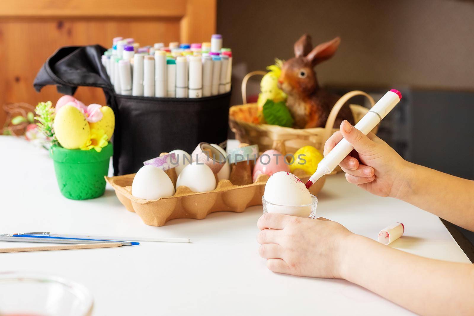 A close-up on the hands of a small Caucasian girl 5 years old paints eggs with special markers paints for the Christian spring holiday of Easter. Girl dressed in a yellow floral dress