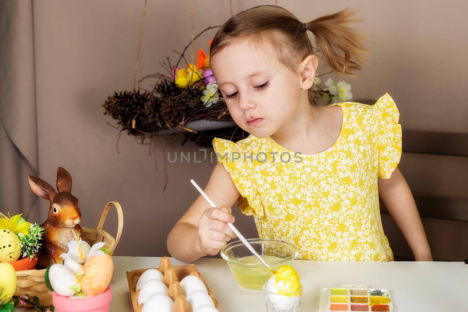 A small Caucasian girl of 5 years old paints eggs with special water paints for the Christian spring holiday of Easter. The girl is dressed in a yellow floral dress and has ponytails.
