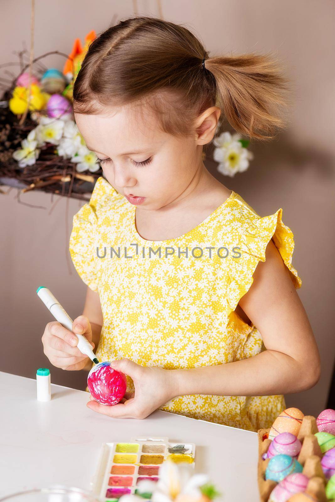 A small Caucasian girl of 5 years old paints eggs with special markers for the Christian spring holiday of Easter. The girl is dressed in a yellow floral dress and has ponytails.