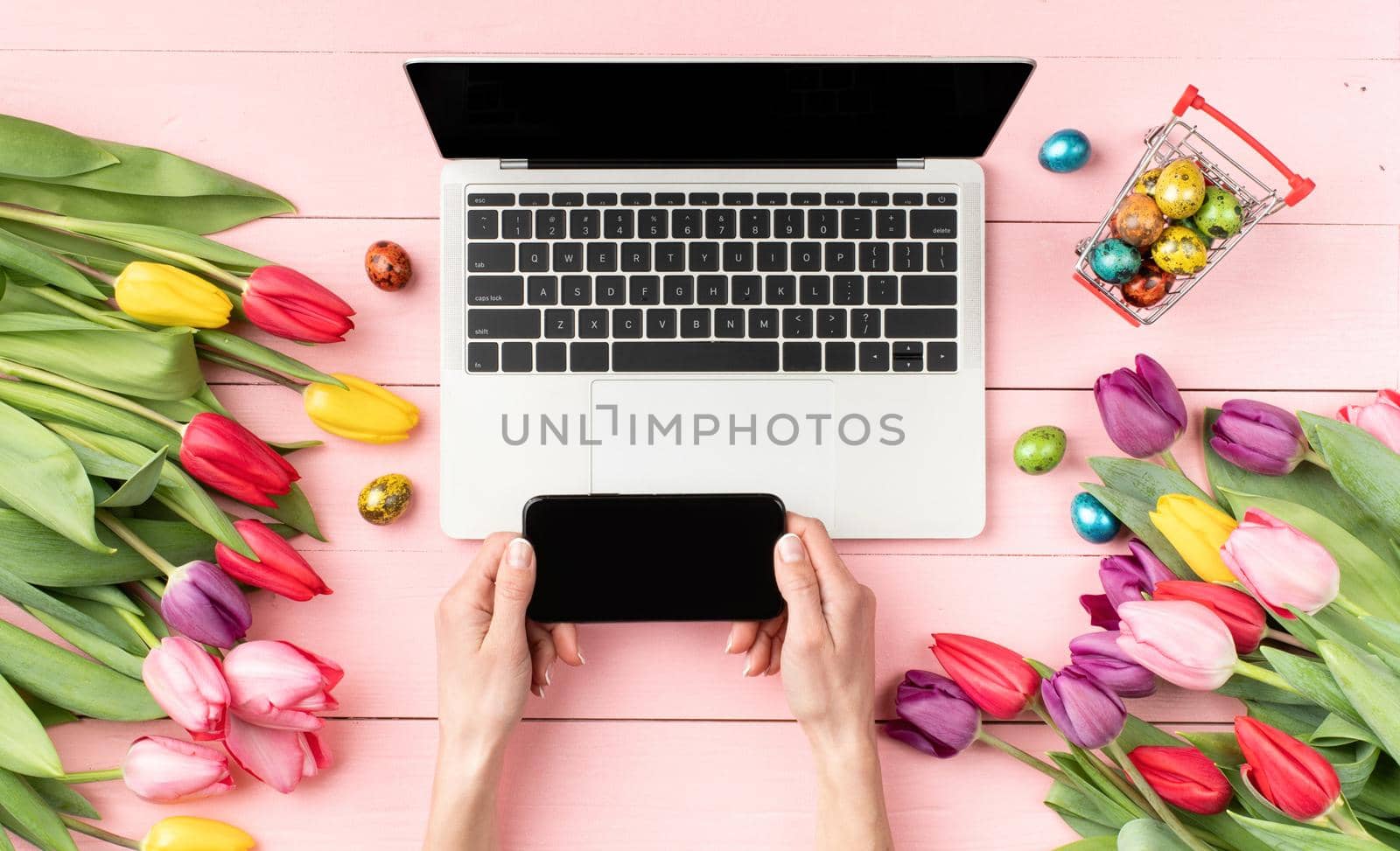 Top view of feminine workspace with laptop computer, tulips, eggs and and female hands using mobile phone on pink background by Desperada