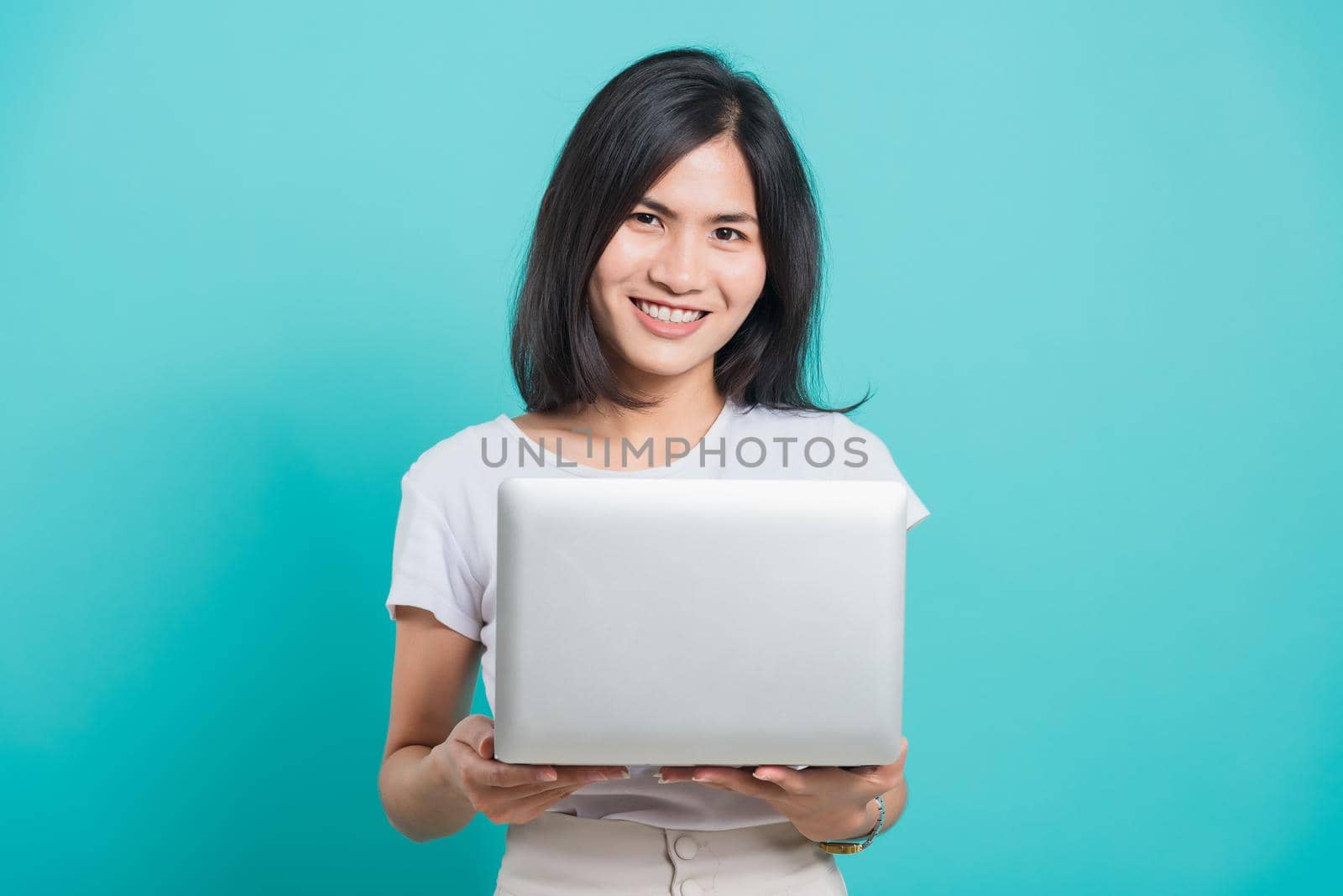 Portrait happy Asian beautiful young woman smile white teeth standing wear white t-shirt, She holding and using a laptop computer, studio shot on blue background with copy space for text