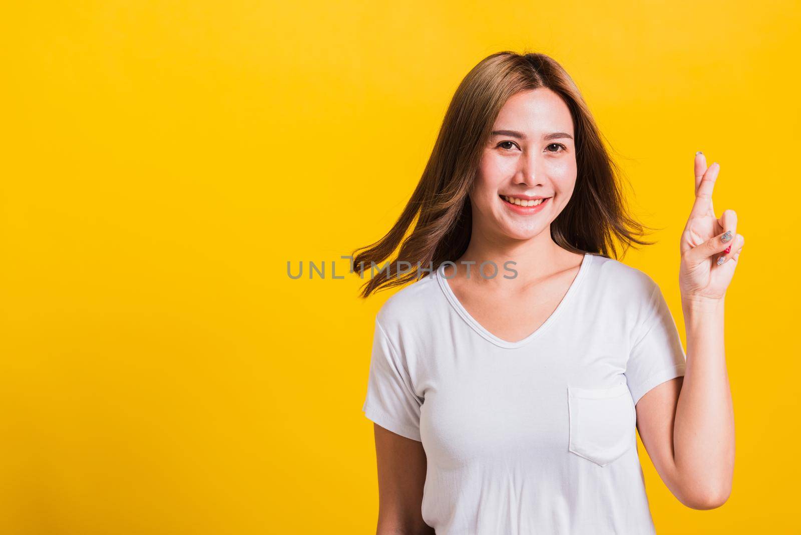 Asian Thai happy portrait beautiful cute young woman smile have superstition her holding fingers crossed for good luck and looking to camera, studio shot isolated on yellow background with copy space