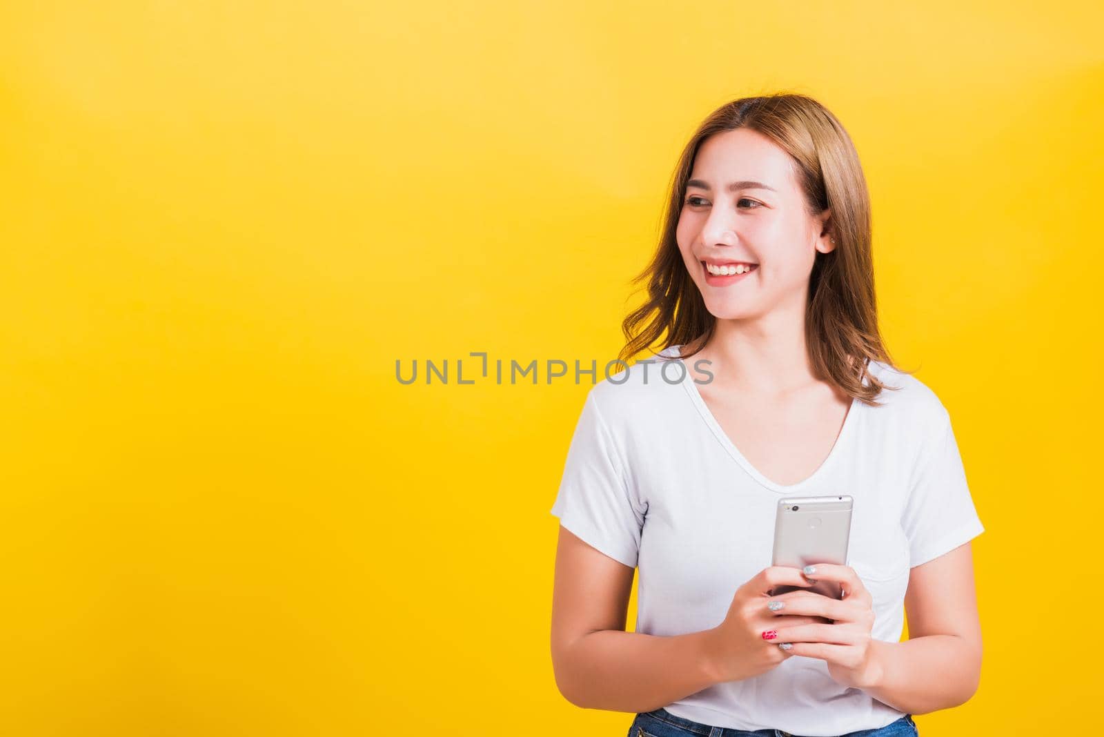 Asian Thai portrait happy beautiful cute young woman smile standing playing game or writing SMS message on smartphone looking to side away space, studio shot isolated on yellow background