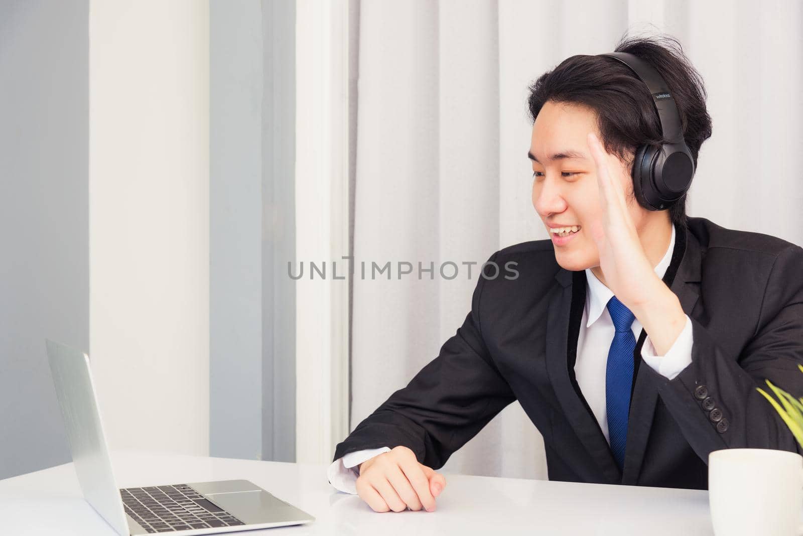 Work from home, Asian young businessman smile wearing headphones and suit video conference call or facetime on desk and raise your hand to say hello greet colleague at home office