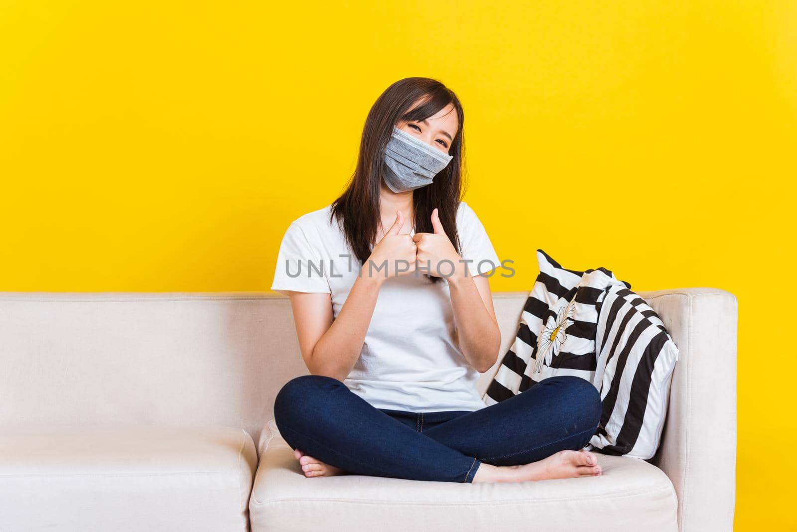 Young woman sitting on sofa wearing medical face mask protective show finger thumb up for good sign by Sorapop