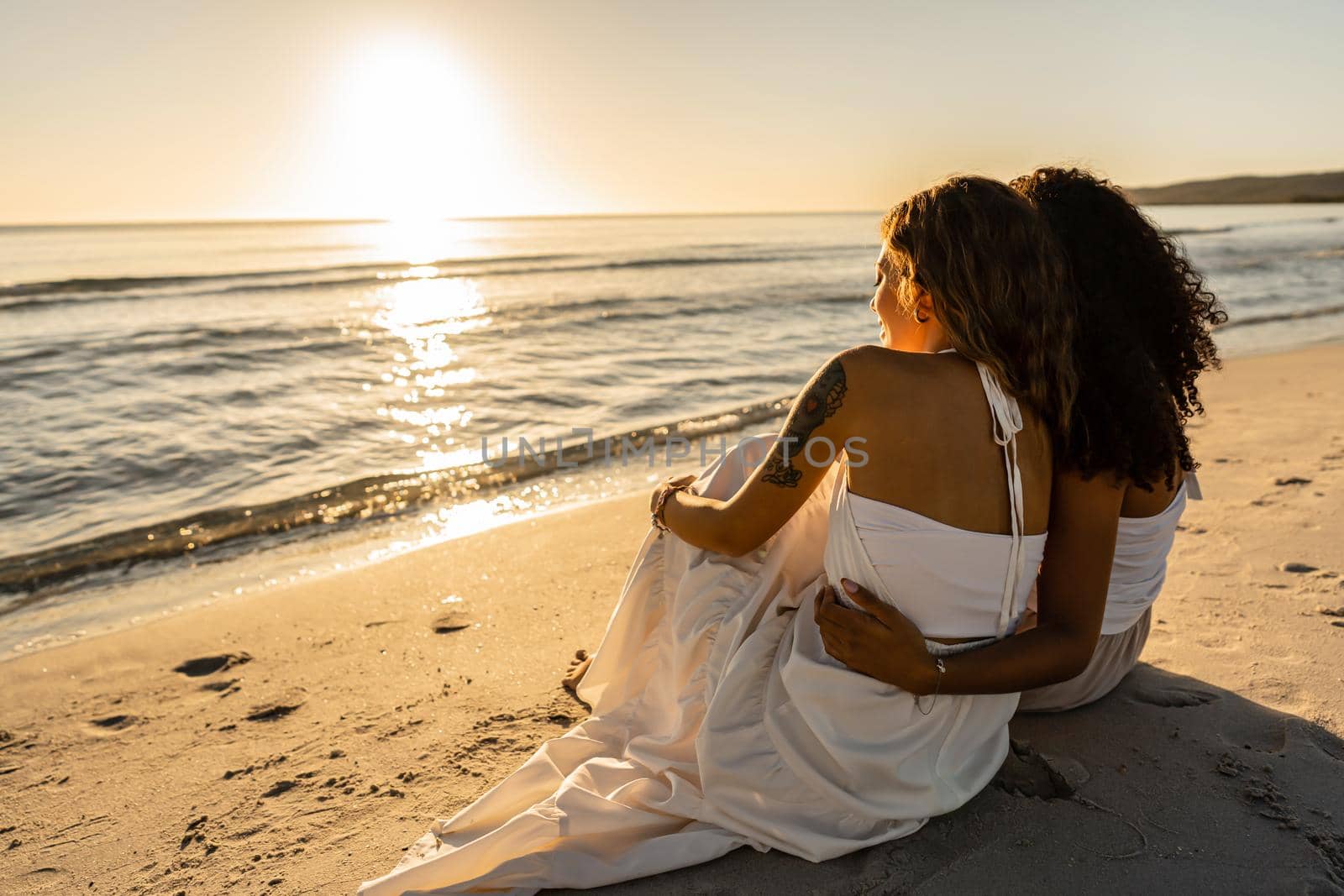 Two lesbian young women view from back sitting on seashore looking at sunset or dawn. Afro american homosexual girl hugs her blonde girlfriend near ocean water in summer vacations. Diversity concept by robbyfontanesi