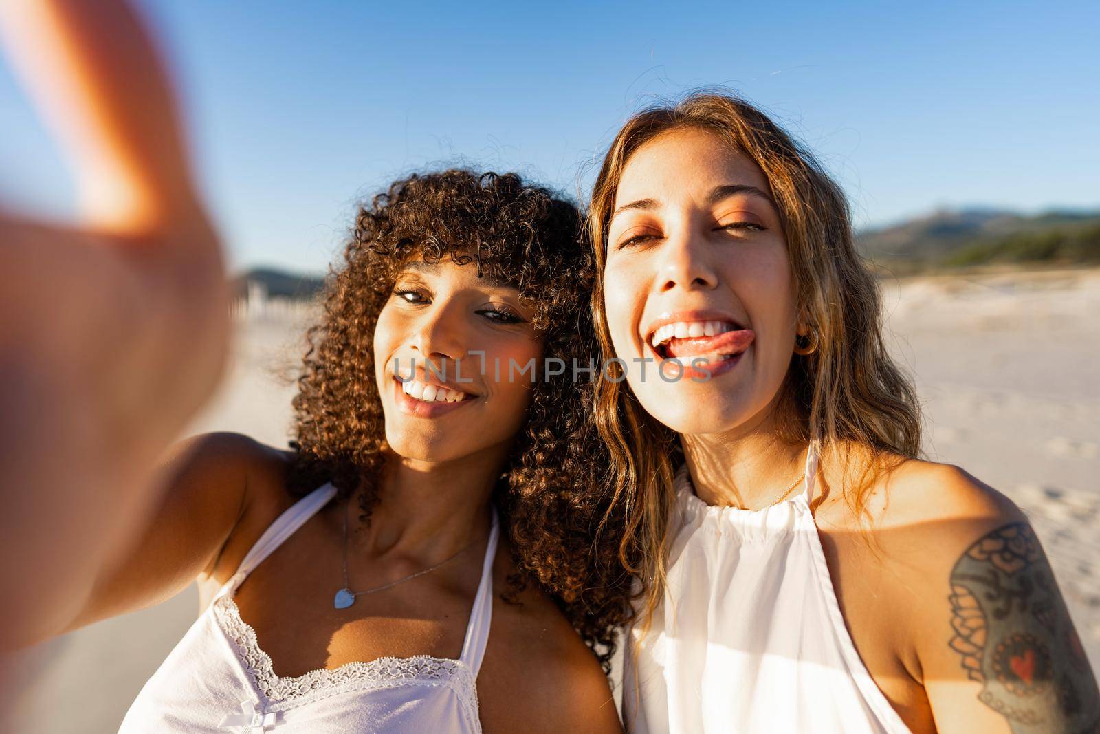 Two happy women multiracial best friends taking selfie on the beach with funny tongue faces wearing boho clothes. Tattooed girl with afro american girlfriend take a funny self portrait in vacations