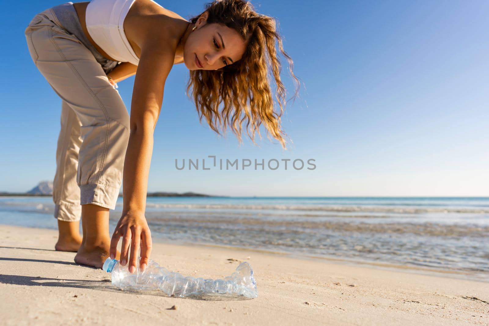 Young woman well educated and sensitive to environmental issues collects an empty plastic bottle abandoned on the seashore to preserve the ocean from plastic waste. Concept of taking care of nature
