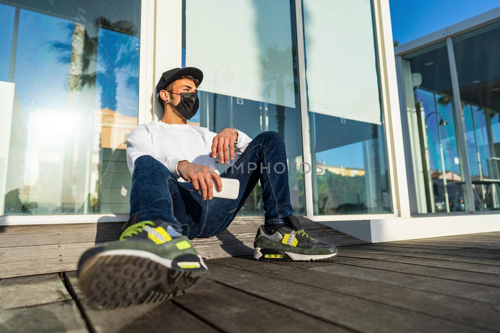 Young modern bored guy sitting on the ground in the city street leaning against a store window wearing Coronavirus protective mask, hat holding smartphone. Concept of new normal young people life