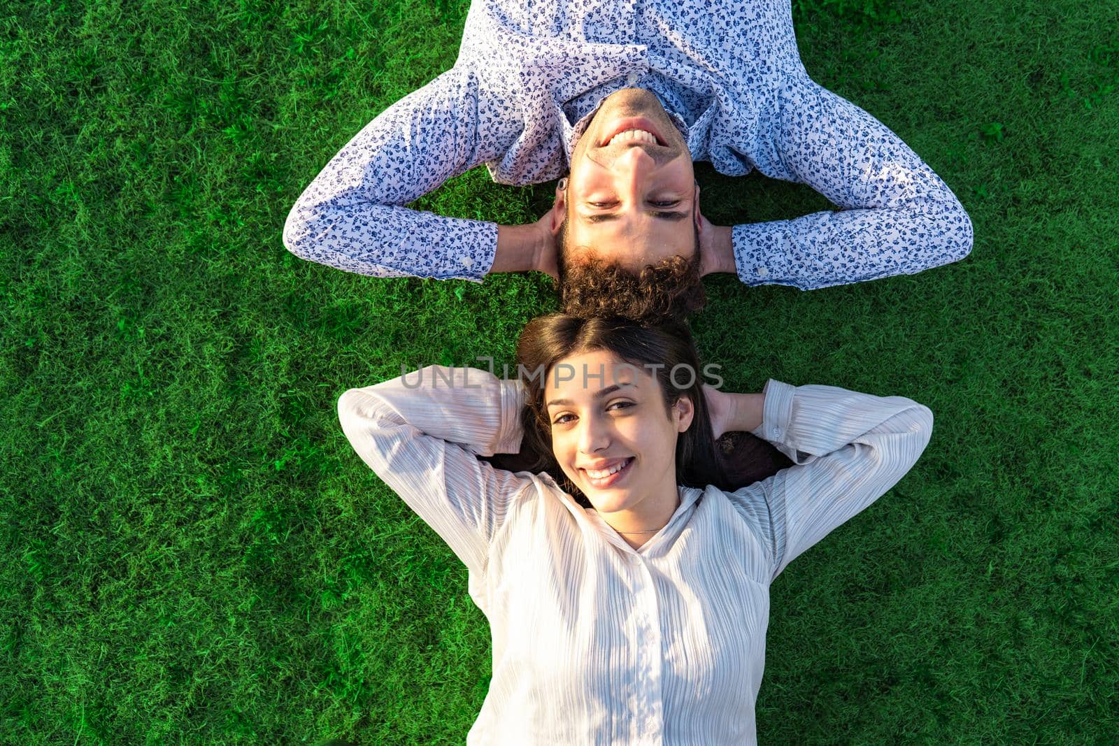 Happy young couple view from top with crossed arms under head smiling looking at camera lying on a green grass meadow with sunset or dawn colored light. Concept of confident people trust in the future