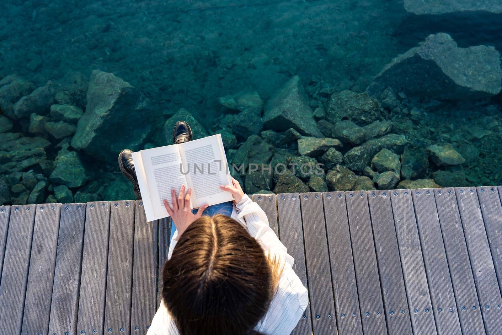 View from top of young school student sitting on a pier on the transparent blue ocean or sea water reading a paper book. Woman relaxing in the nature spends time with her favorite romance novels