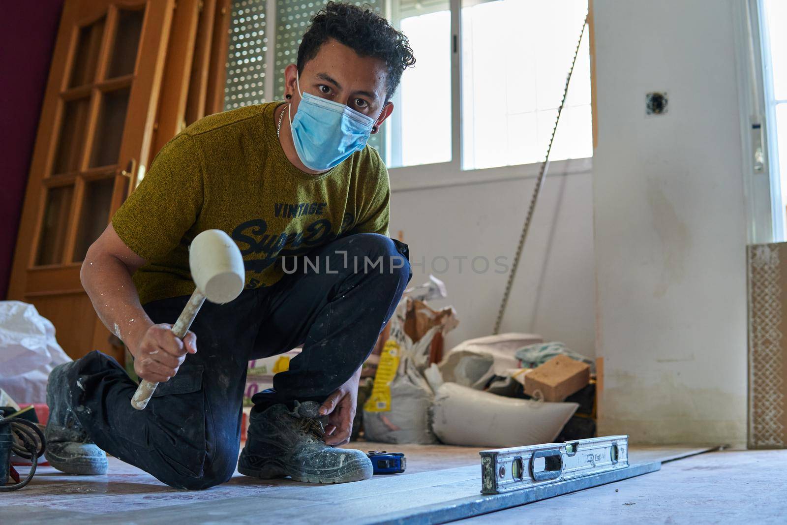 Mason tiling a room with a mustard-colored t-shirt and rubber pot and level and with a mask