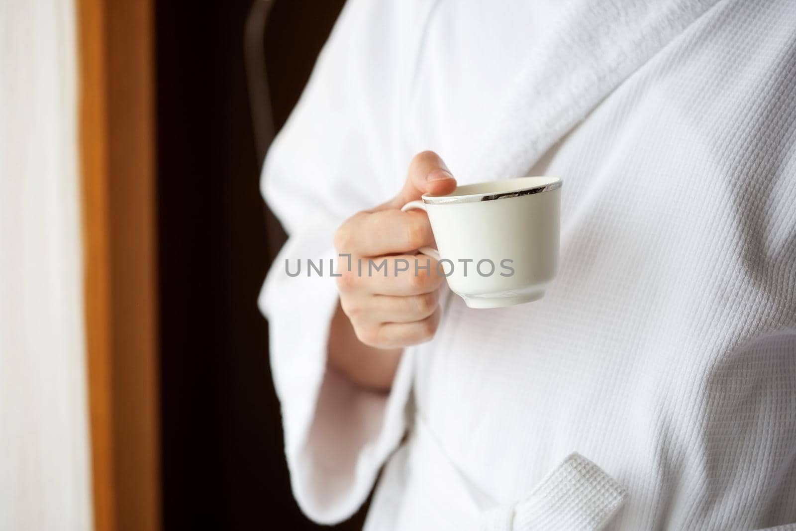 A man in a white terry dressing gown holds a coffee mug in front of a window. by galinasharapova