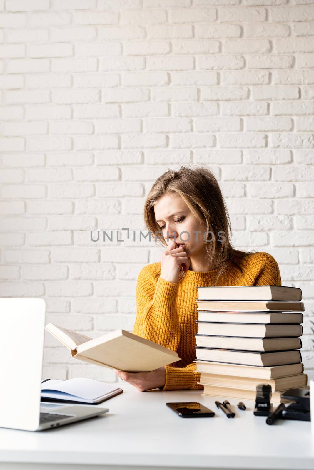 Young thoughtful woman in yellow sweater studying using laptop and reading a book