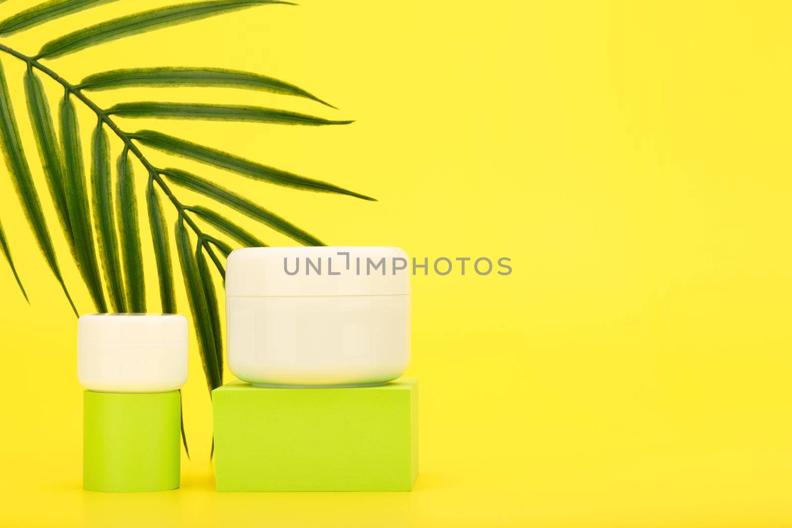 Cream or mask and lip balm or under eye cream on green podiums against yellow background with palm leaf and copy space by Senorina_Irina