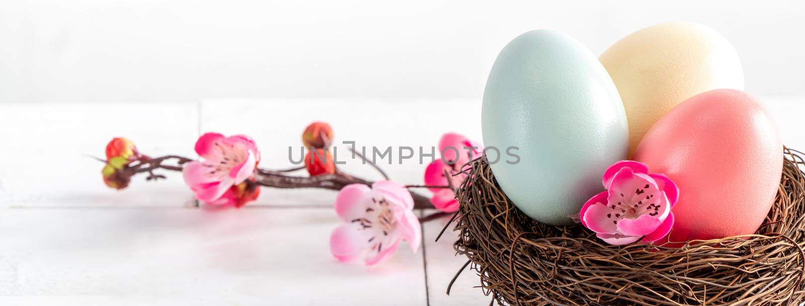 Close up of colorful Easter eggs in the nest with pink plum flower on bright white wooden table background.