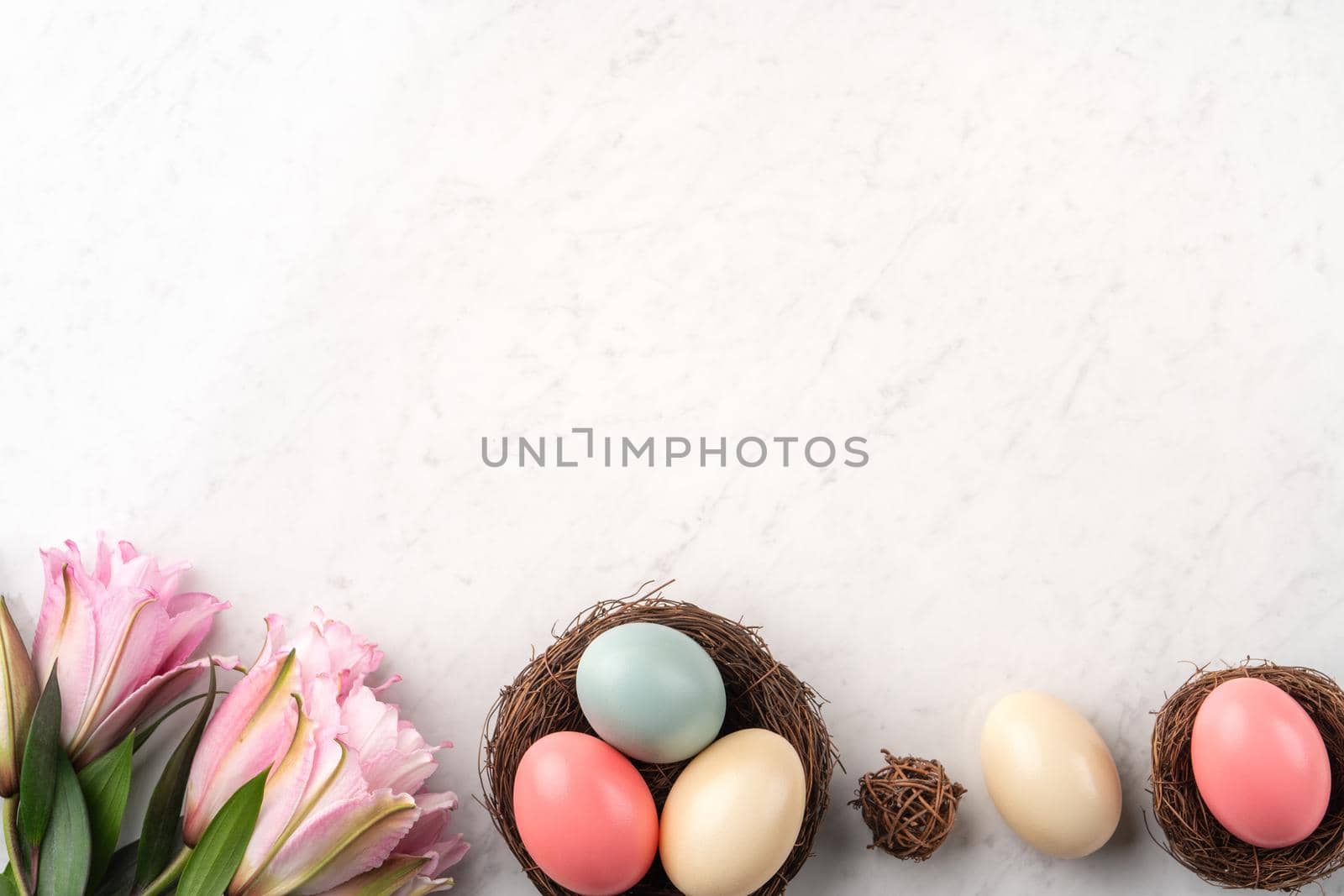 Colorful Easter eggs in the nest with pink lily flower on bright marble white table background.