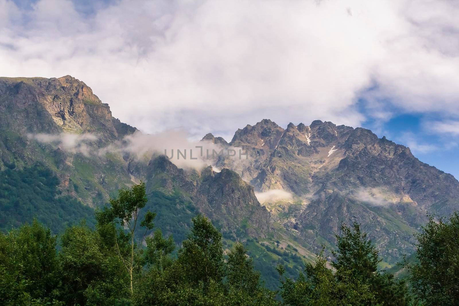 High mountains of the Caucasus with a beautiful view. Green vegetation and shrubs on sharp rocks above a blue sky. Magnificent mesmerizing landscape.