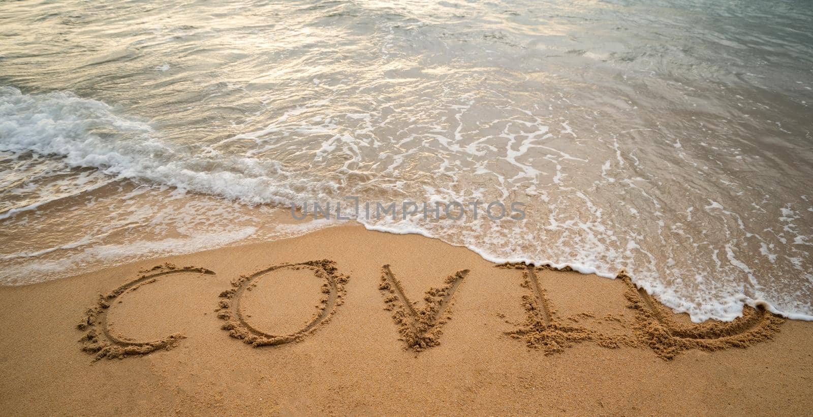 Handwriting COVID on sand and foam wave on beach. Coronavirus concept. by sirawit99