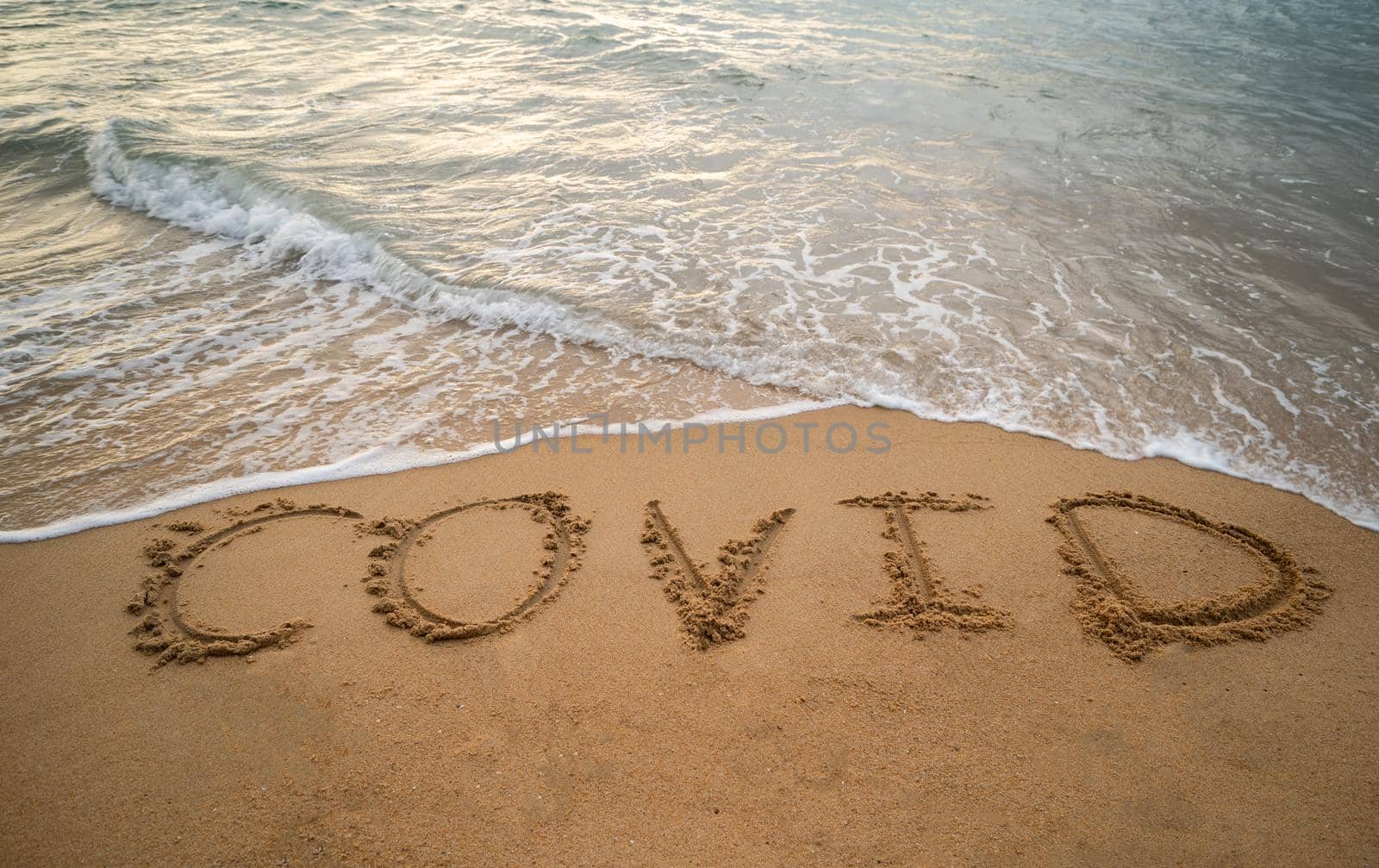 Handwriting COVID on sand and foam wave on beach. Coronavirus concept. by sirawit99