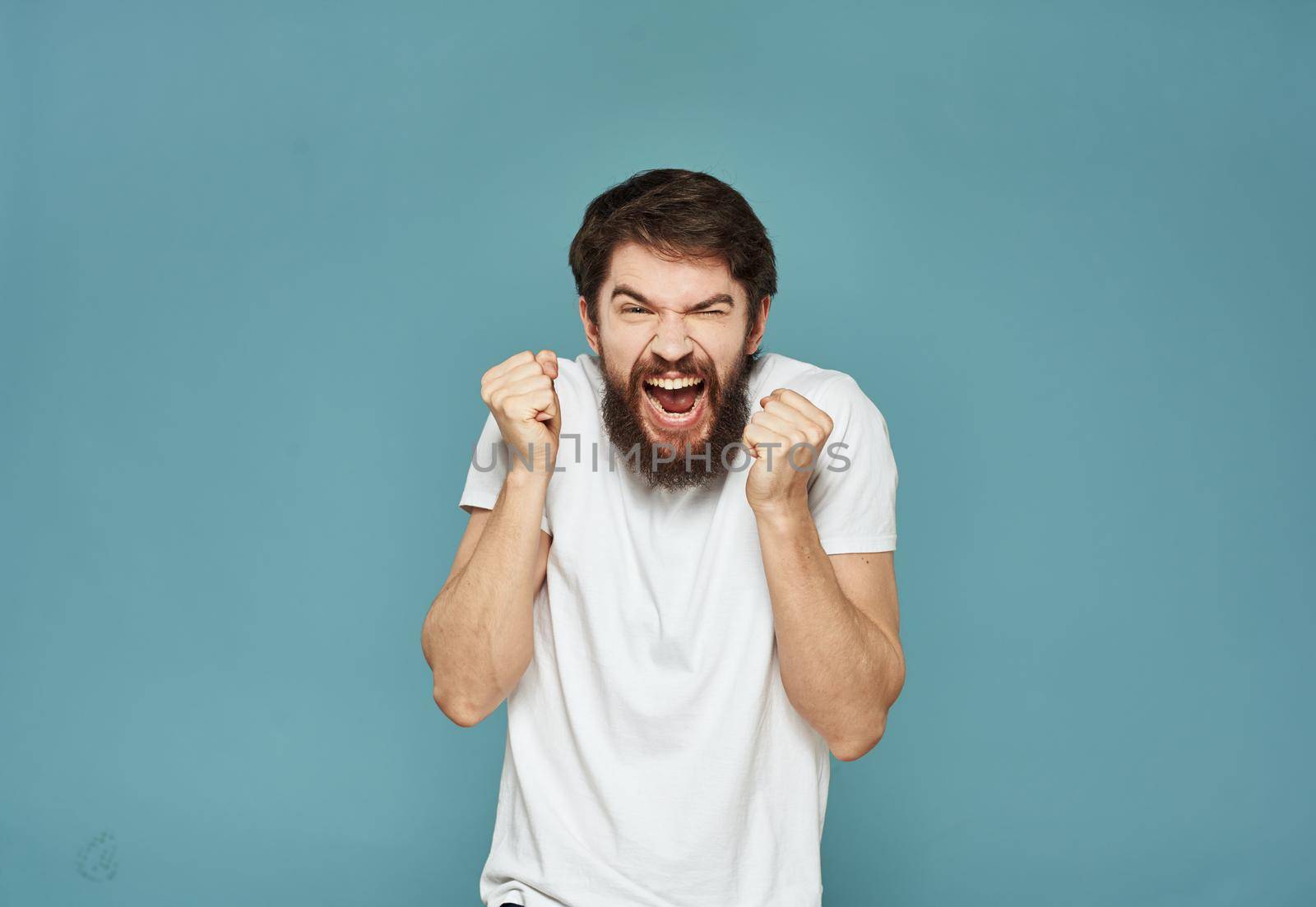 A successful man in a white T-shirt clenched his hands into a fist on a blue background by SHOTPRIME