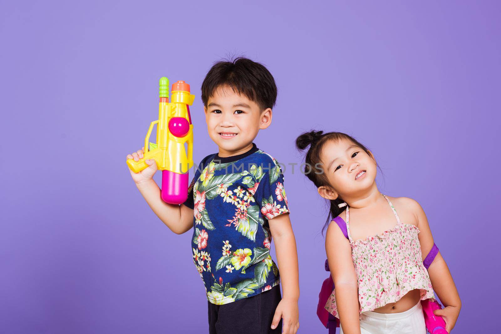 Two Happy Asian little boy and girl holding plastic water gun, Thai children funny hold toy water pistol and smile, studio shot isolated on purple background, Thailand Songkran festival day culture.