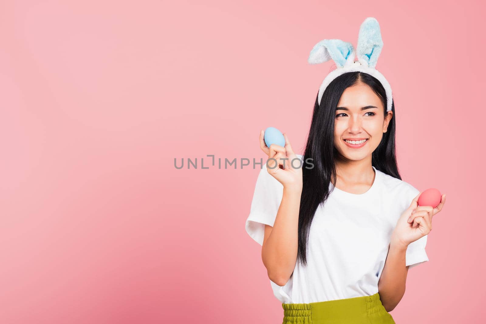 Happy Easter concept. Beautiful young woman smiling wearing rabbit ears holding colorful Easter eggs gift on hands, Portrait female looking at side away, studio shot isolated on pink background