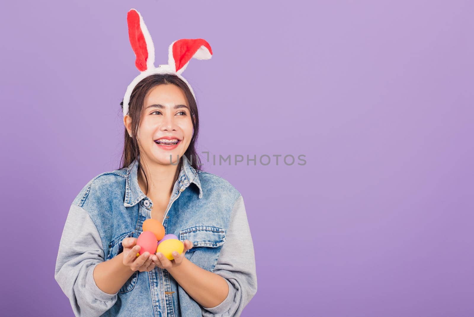 Happy Easter concept. Beautiful young woman smiling wearing rabbit ears and denims hold colorful Easter eggs gift on hands, Portrait female looking at side, studio shot isolated on purple background