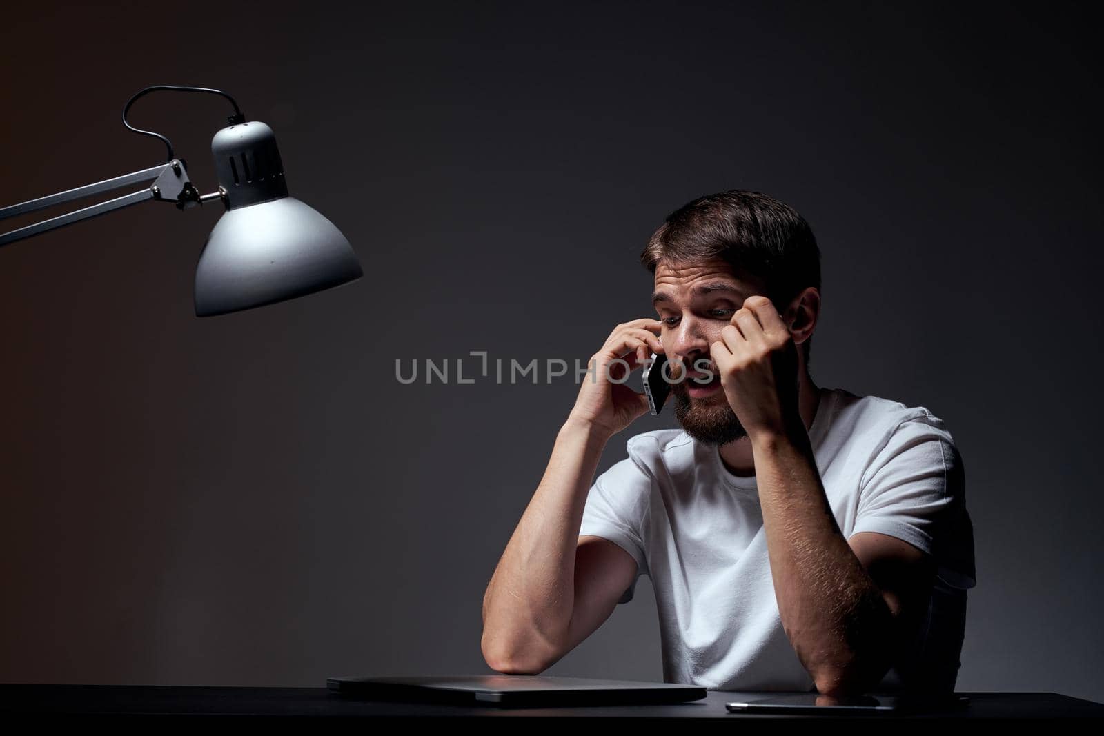 a man in a white t-shirt at his desk laptop lamp dark background. High quality photo