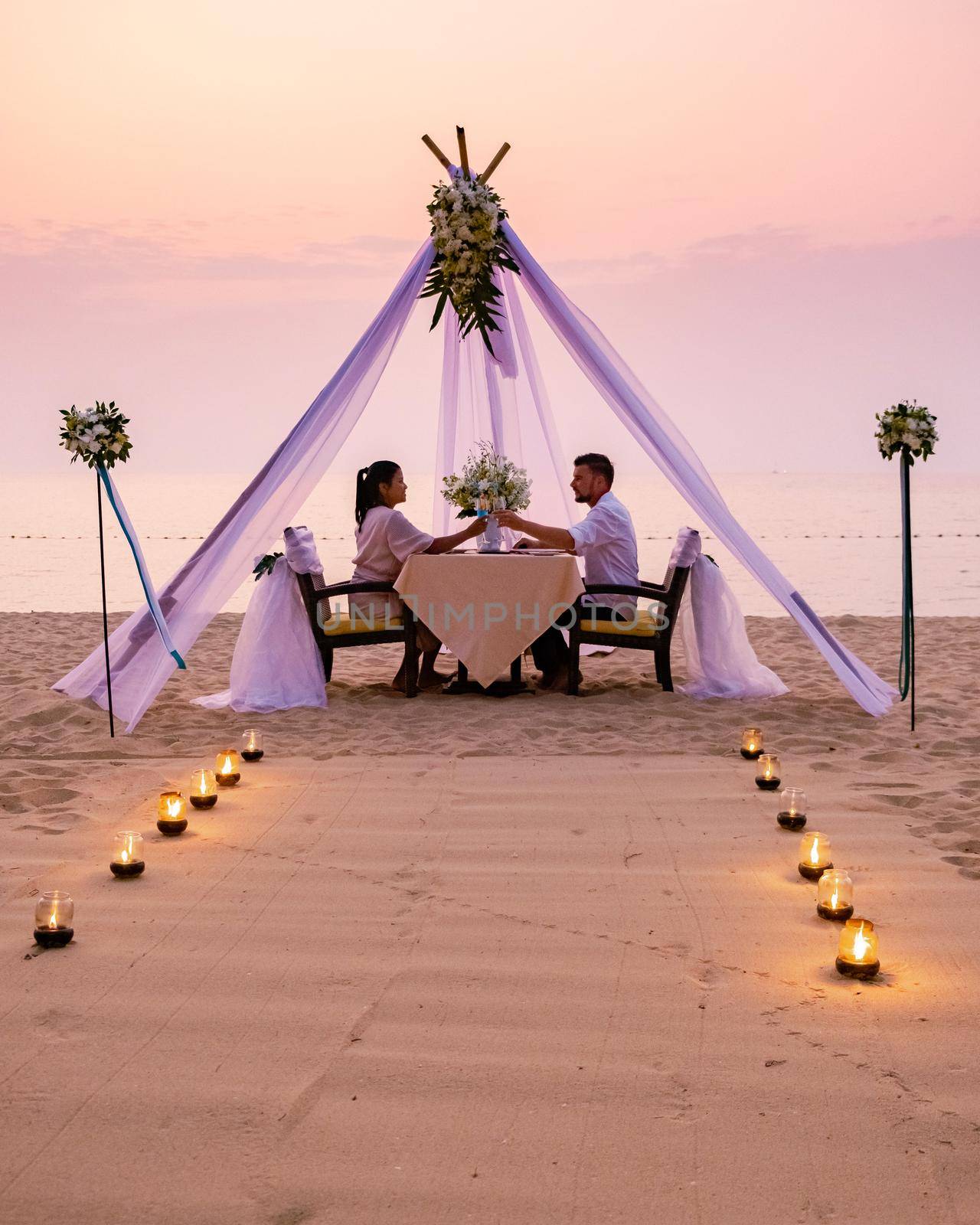 Loving couple share a romantic dinner with candles, lanterns and wine glasses at sea beach sand against wonderful sunset by fokkebok