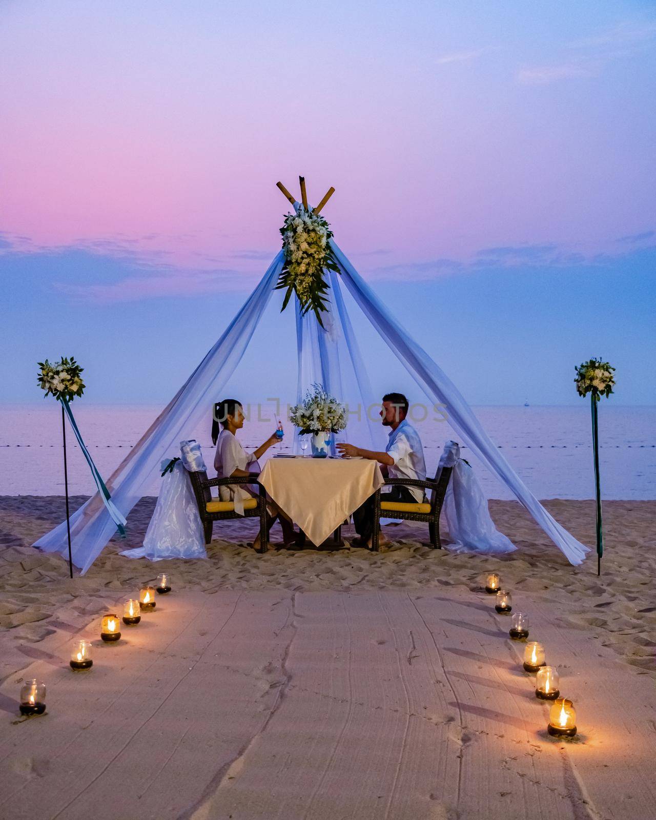 Loving couple share a romantic dinner with candles, lanterns and wine glasses at sea beach sand against wonderful sunset by fokkebok