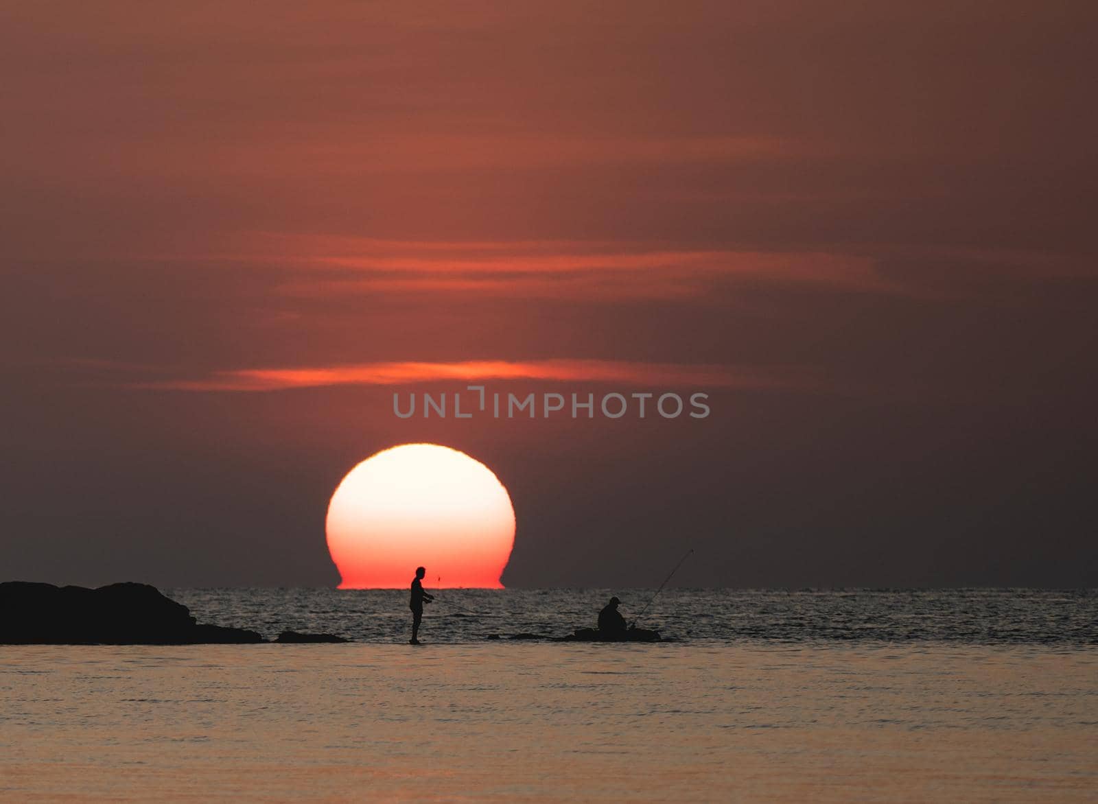 Sunset ocean view. Sun above the sea on orange sky. Fisherman with fishing rod on the rock.