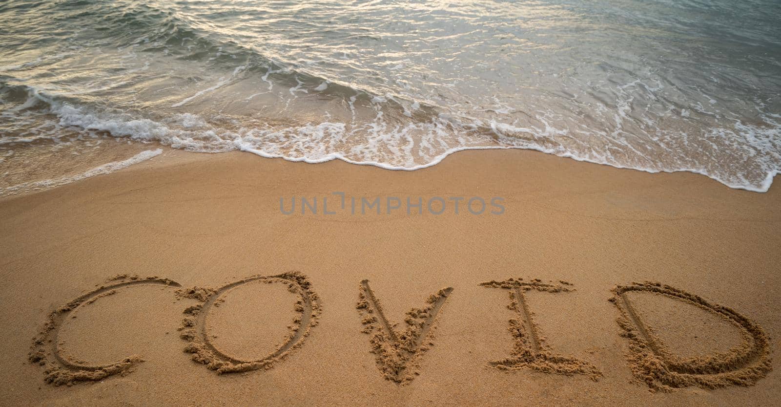 Handwriting COVID on sand and foam wave on beach. Coronavirus concept.