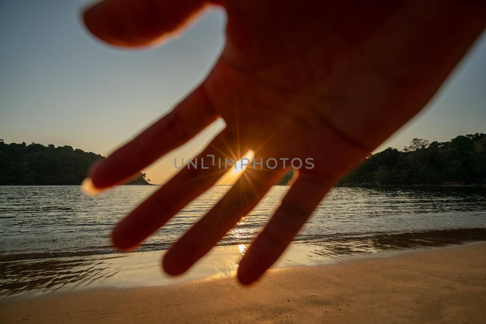 Man hand up front to stop camera. Sunset ocean view. Sun above the sea on orange sky.