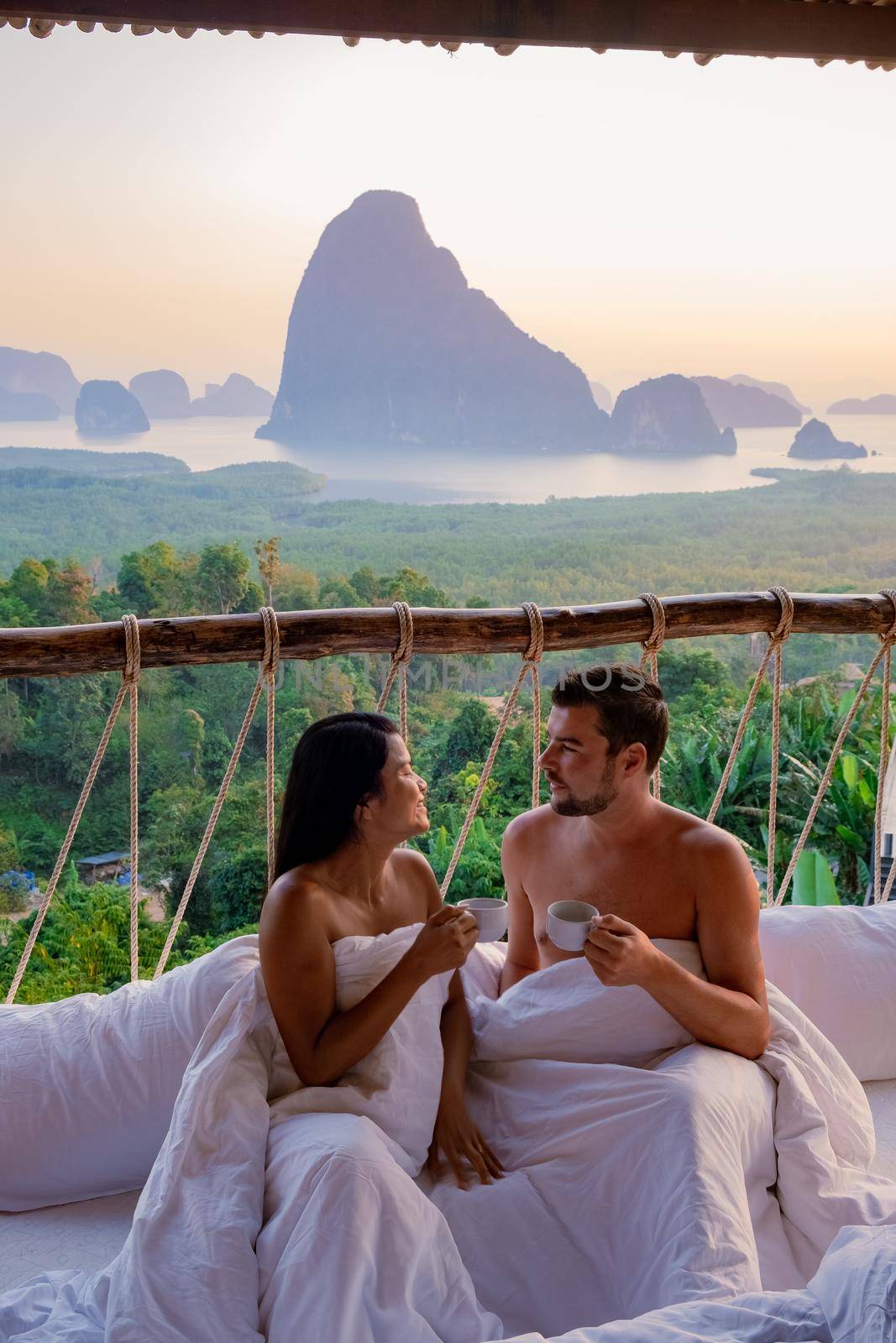 Phangnga Bay, couple waking up in bed in nature jungle looking out over ocean and jungle during sunrise at the wooden hut in the mountains of Thailand men and woman mid age morning sunrise Phanga Thailand