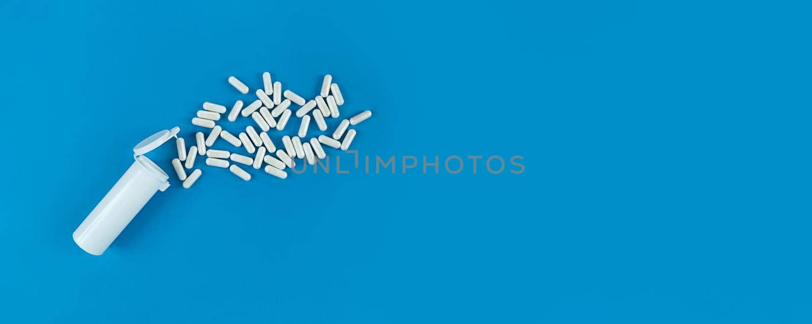 White plastik medical bottle and scattered pills capsules on a blue backdrop. Banner with copy space.